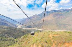 PANACHI – Chicamocha National Park A cable car travels high above a lush, mountainous valley with a river winding through the landscape under a partly cloudy sky.