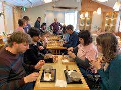 A diverse group of people, including adults and children, participate in a matcha-making experience, gathered around wooden tables with tea bowls and bamboo whisks.