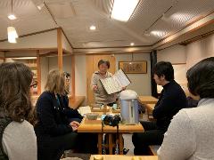 A group of people gather around a table as a woman presents information from an open book, with a rice cooker and tea ceremony items visible.