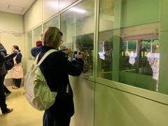 A woman wearing a black coat and a large white bag over her shoulder takes a photo of a matcha-making machine through a glass partition.