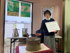 A woman presents a sign with questions about matcha powder amounts while standing in front of a tea grinding stone and display about tea.