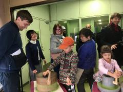 A group of people, including children, are gathered around stone mills, some actively grinding, in what appears to be a matcha-making experience.