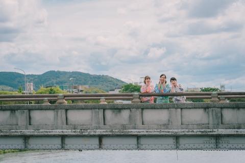 Three women in colorful kimonos pose on a bridge with hands on their faces, set against a backdrop of a cloudy sky and distant mountains.