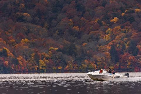 A boat with three people cruises on a lake with a hillside covered in autumn foliage in the background.