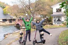 A man and woman cheerfully pose with their electric scooters beside a lake in a scenic rural setting.
