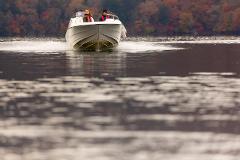 A speed boat carries three people across a calm lake, with the blurry, autumnal colors of the distant shore serving as a backdrop.