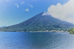 A serene lake reflects a vast, lush green mountain under a bright blue sky with scattered clouds.