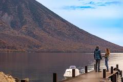 A couple stands on a dock overlooking a tranquil lake with a tree-covered mountain in the background.