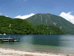 A tranquil lake is framed by lush green mountains and a large, imposing peak under a bright blue sky.