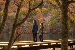 A couple stands on a wooden walkway overlooking a lake surrounded by autumn foliage.