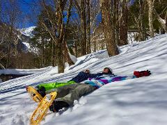 Two people in snow gear relax on a snow-covered slope in a sunny forest with snow-capped mountains in the background.