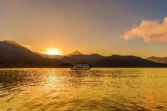A ferry glides across a shimmering golden lake as the sun sets behind silhouetted mountains under a warm sky.