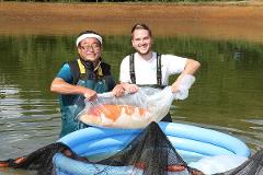 Professional breeder-guided Koi farm tour in the sanctuary of Nishikigoi Two men hold a large koi fish in a plastic bag over a blue inflatable pool in a pond.