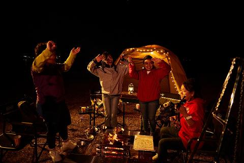 A group of four Asian women enjoy a barbecue by a campfire at night near a tent decorated with string lights.