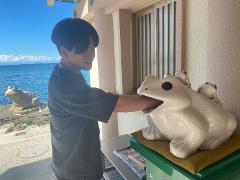 A person reaches into the mouth of a large white frog statue with smaller frogs on its back, with a blue sea and sky in the background.