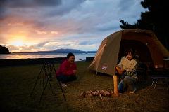 Two women are camping by the sea at sunset, one laughing and the other preparing firewood near a Coleman tent.
