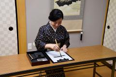 A smiling woman in a kimono practices Japanese calligraphy, holding a brush over a sheet of white paper with inked characters.