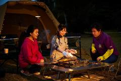 Three young women are camping and starting a fire in a metal fire pit while crouching around it.