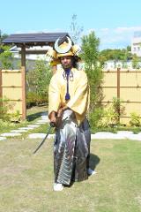 一名打扮成武士的男子站在花园里，手持武士刀，身穿黄色和服，头戴传统头盔。