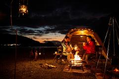 Two women enjoy a campfire at a campsite near the beach with a tent and lanterns illuminating the scene under a dusky sky.