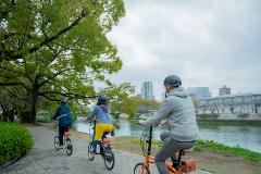 Three people are riding their bikes in a line down a paved path beside a body of water with trees and buildings in the background.