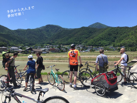 Nagano Onsen Town Cycling Tour with Ice Cream – Half Day Ride A group of people with bicycles and a child carrier stand by a road with rice paddies and mountains in the background.