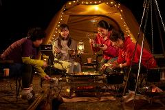Four people are gathered around a grill table with a campfire, eating and cooking outdoors, illuminated by lanterns and string lights around a tent.