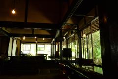 A dimly lit traditional Japanese building interior with dark wooden beams and posts, featuring large windows that reveal lush green foliage outside.