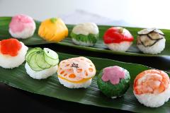 A colorful assortment of temari sushi rests on two green banana leaves, featuring toppings like shrimp, cucumber, lotus root, and various other vibrant ingredients.