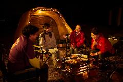 Four people are sitting around a grill with a lit fire, cooking food in front of a Coleman tent at night, all laughing and having a good time.