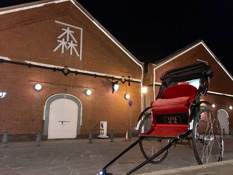 Private Night Rickshaw Adventure in Hakodate Bay Area A rickshaw with a red cushioned seat is parked in front of a brick building decorated with lights at night.