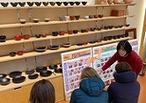 Experience 200 Years of Craftsmanship: Wajima Lacquerware Studio Tour (Visit a craftsman's home) A woman points to a display of traditional Japanese lacquerware bowls on shelves and charts for a group of visitors in a studio.