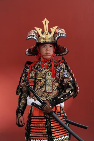 一名身穿华丽武士盔甲（包括精致的头盔）的男子在红色背景下手持两把剑。
