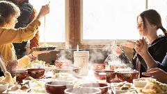 A group of people enjoy a meal of steaming noodles and tempura at a table laden with dishes, set near a window with natural light.
