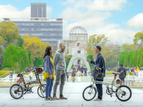 Two tourists with their folding bikes listen to a local at the Hiroshima Peace Memorial, with the iconic Atomic Bomb Dome and Memorial Museum in the background.