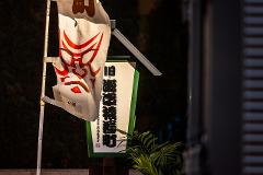 A flag with a red oni mask design hangs beside a sign with Japanese characters, in front of a dimly lit background with some foliage.