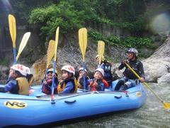 A group of children and an adult are rafting down a river, all wearing helmets and life jackets.