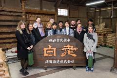 A group of tourists poses in front of a wooden sign at a Japanese brewery, some holding shopping bags.