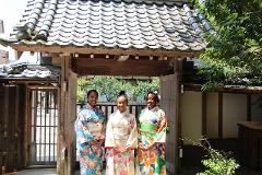 Three women wearing colorful kimonos stand in front of a traditional Japanese gate with a tiled roof on a sunny day.