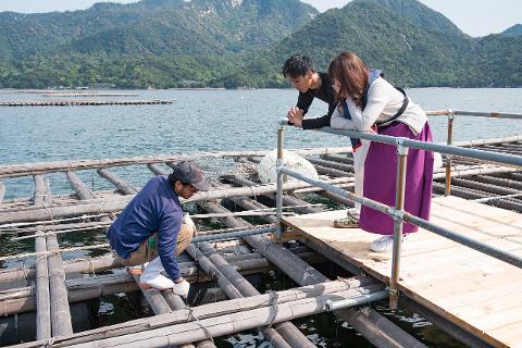 Üç kişi, arka planda dağlar varken suya bakan ahşap bir platformun üzerinde duruyor, bir kişi çömelmişken, diğer ikisi korkuluğa yaslanmış dışarıyı izliyor.