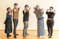 A group of people with traditional Japanese Kabuki makeup and costumes pose with their hands extended on a wooden floor.