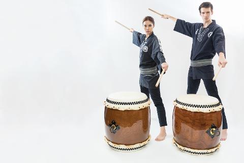 Two performers in traditional Japanese attire with taiko drumsticks stand next to taiko drums against a white background.
