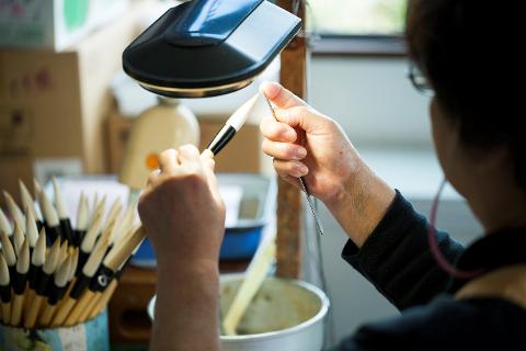 A person is meticulously using a metal tool to shape the tip of a brush, illuminated by a bright lamp, with many similar brushes in the background.