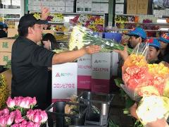 At a flower market, a man in a black shirt and cap gestures while handing a bundle of white flowers to a group of customers wearing blue hats. Pink roses and boxes labeled "春田フラワー コロンビアの花" are visible.