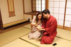 A happy couple in traditional Japanese attire, kneeling on tatami mats, holding tea bowls in a serene room with shoji screens.