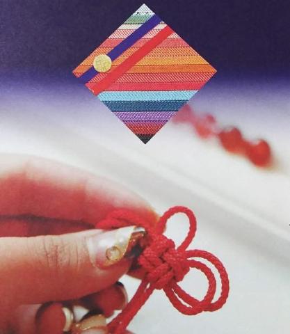 Um close mostra uma mão segurando um nó decorativo vermelho, com um desenho colorido em forma de diamante acima e contas vermelhas desfocadas no fundo.