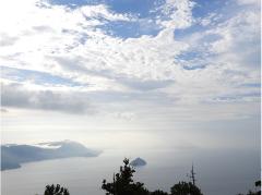 A scenic view of a lake surrounded by mountains under a cloudy sky, partially obscured by fog, with trees in the foreground.