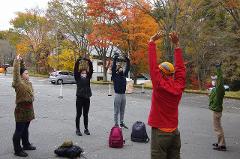 Embrace Beautiful Mother Nature on an E-Bike Tour of Yatsugatake A group of people wearing masks and gloves practice exercises outdoors with colorful autumn trees in the background.