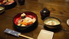 A wooden table is set with a bowl of assorted sushi, miso soup, a side dish, chopsticks, and a small square plate, ready for a meal.