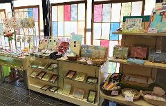 A variety of Japanese paper crafts, including fans, notepads, and framed artwork, are displayed in a shop with stained glass windows in the background.
